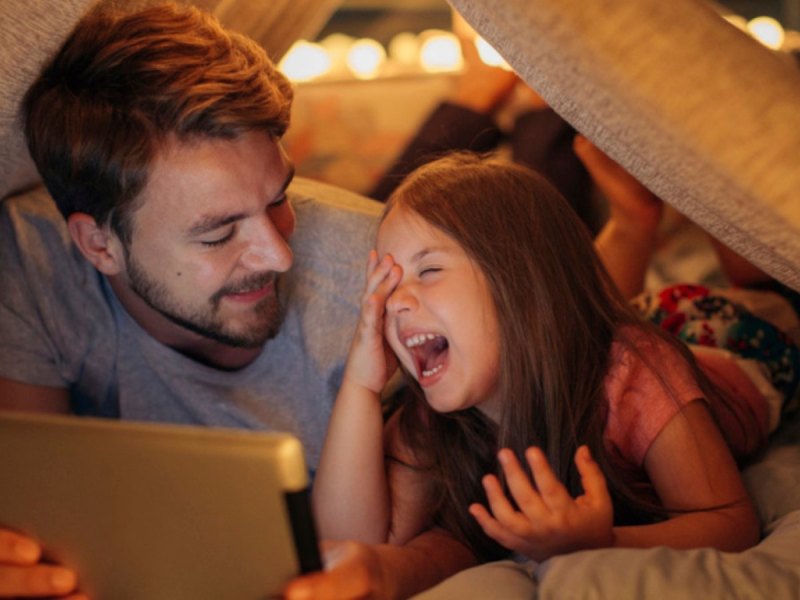 Vater und Kind liegen im Bett und schauen auf ein Tablet.