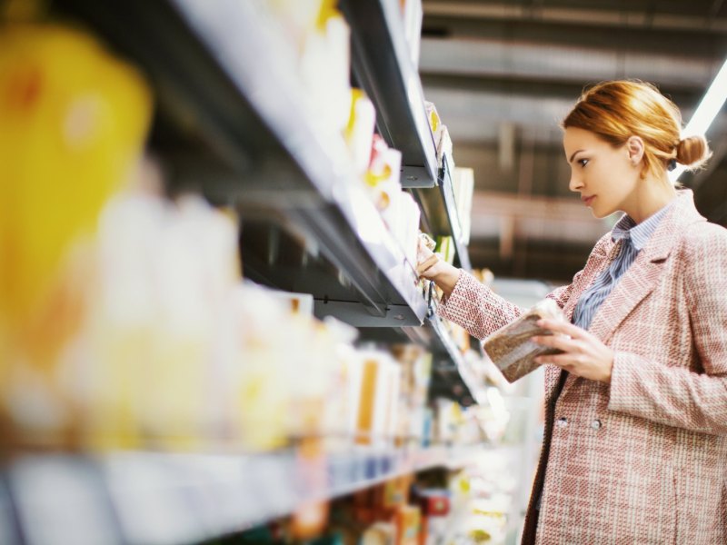 Junge Frau steht vor einem Regal im Supermarkt und hält eine Packung Vollkornbrot in der Hand.