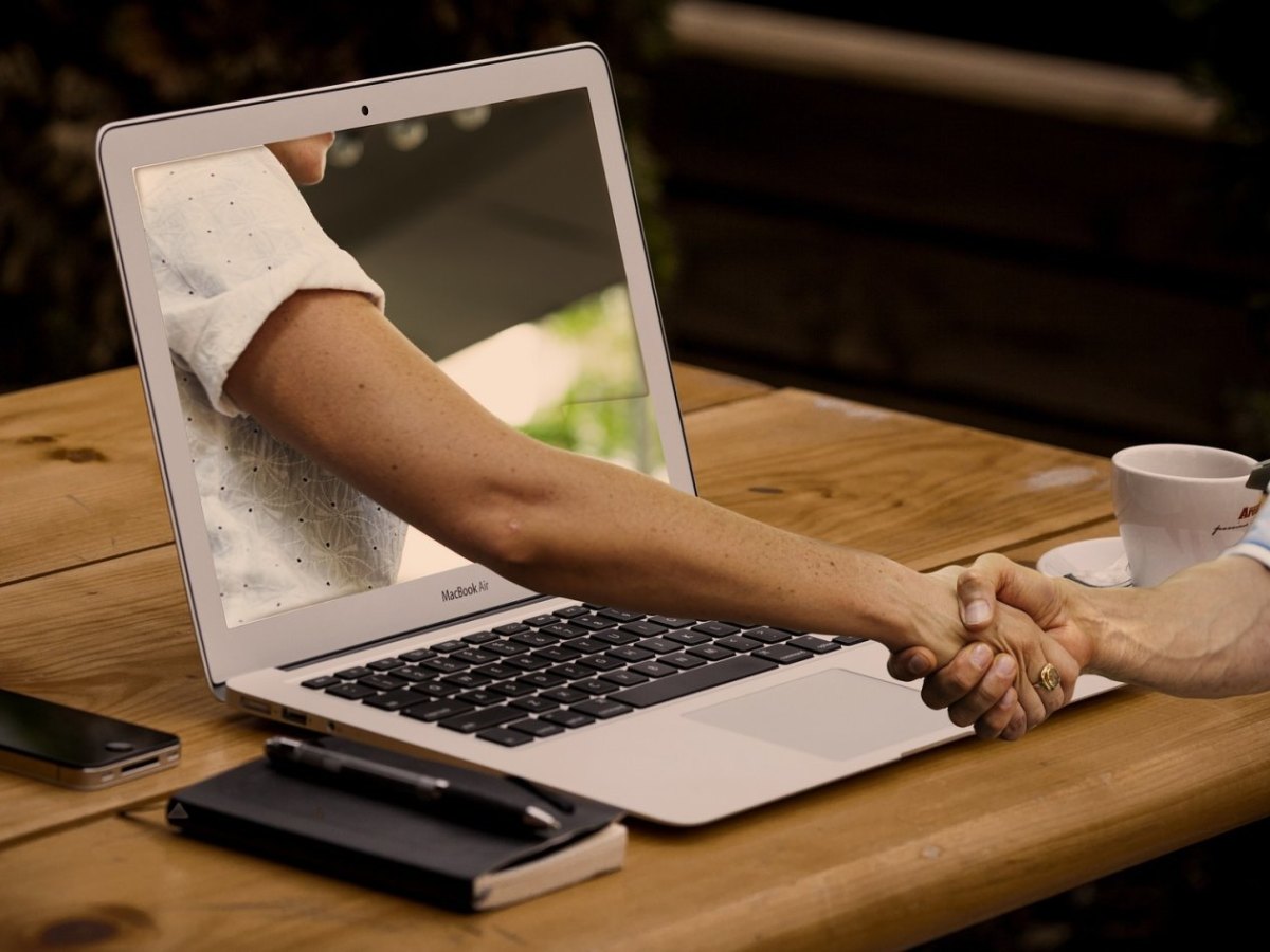 Eine Hand reicht einer anderen durch einen Computer hindurch die Hand.