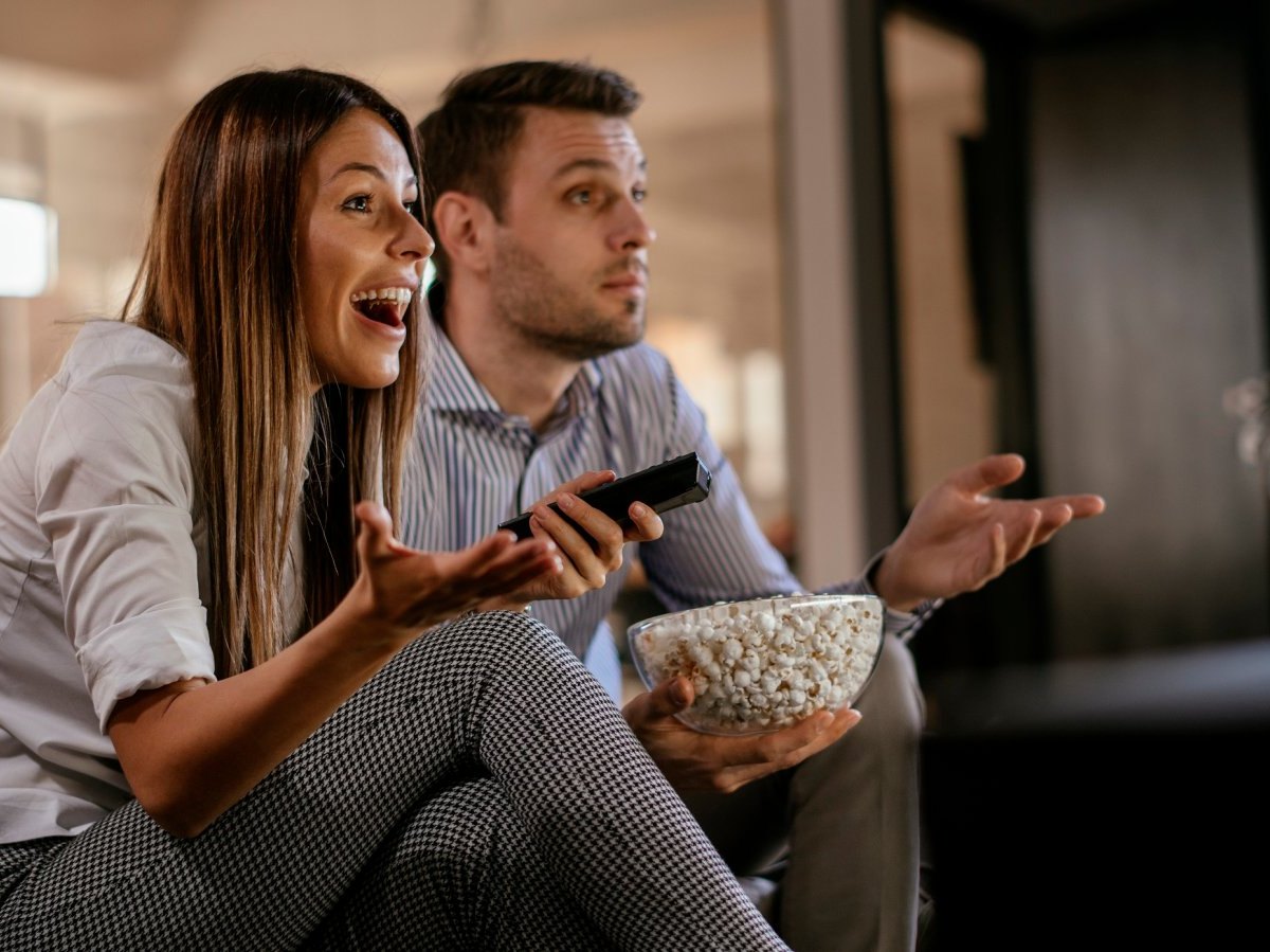 Paar sitzt überrascht und glücklich und mit einer großen Schüssel Popcorn vor dem Fernseher.