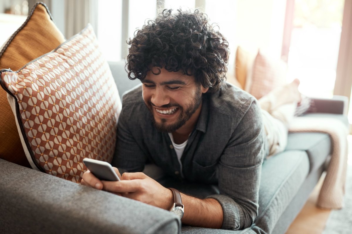 Mann liegt gemütlich auf dem Sofa und blickt glücklich auf sein Handy.