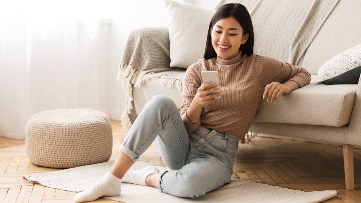 Frau lächelnd am Handy an eine Couch gelehnt.