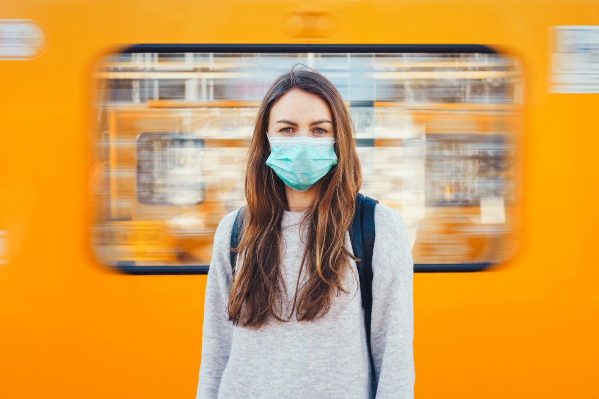 Frau mit Mundschutz steht vor einer vorbeifahrenden Bahn.