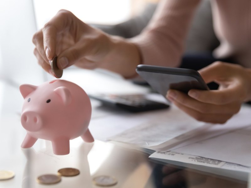 Frau hält Handy in der Hand und schmeißt Geld in ein Sparschweinchen.