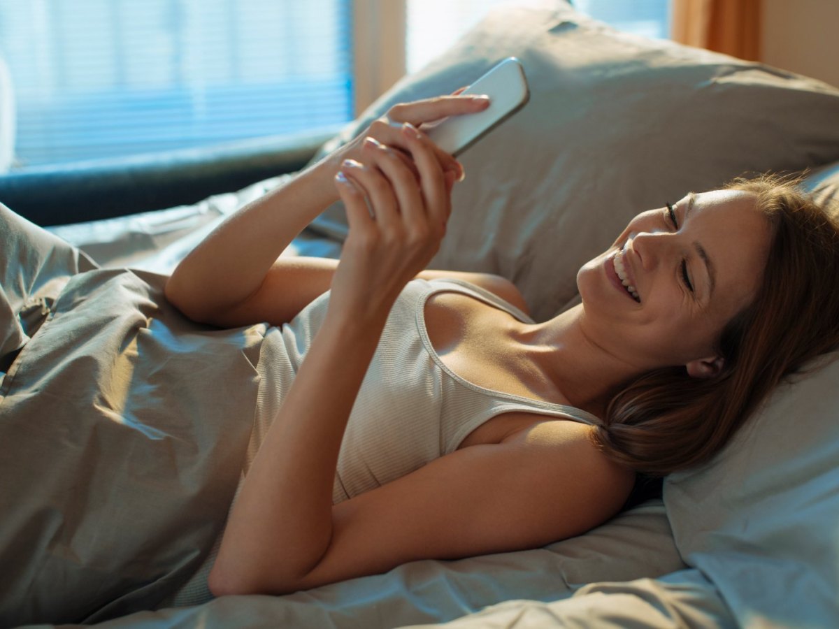 Frau liegt lachend im Bett und schaut auf ihr Handy.