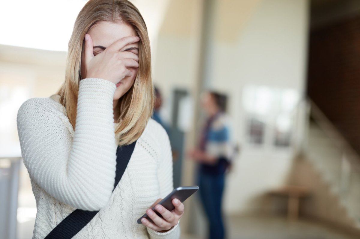 Eine Frau verdeckt peinlich berührt ihr Gesicht mit der Hand und hält das Handy in der anderen Hand.