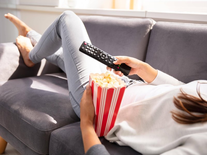 Person auf Couch mit Popcorn und Fernbedienung