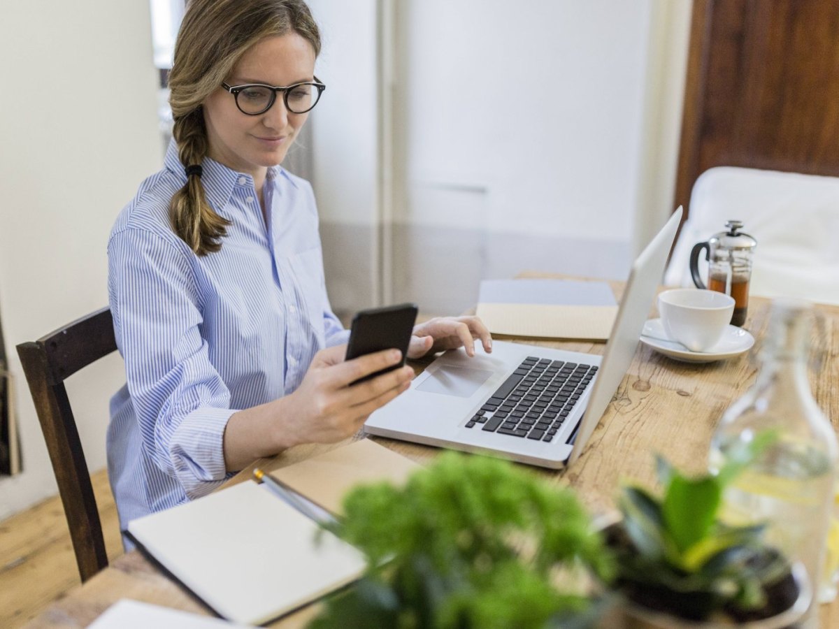 Frau sitzt am Laptop und schaut auf ihr Smartphone