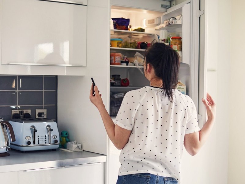 Frau mit Handy schaut in den Kühlschrank.