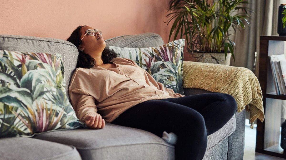 Frau sitzt nachdenklich und geschafft auf dem Sofa und blickt nach oben.