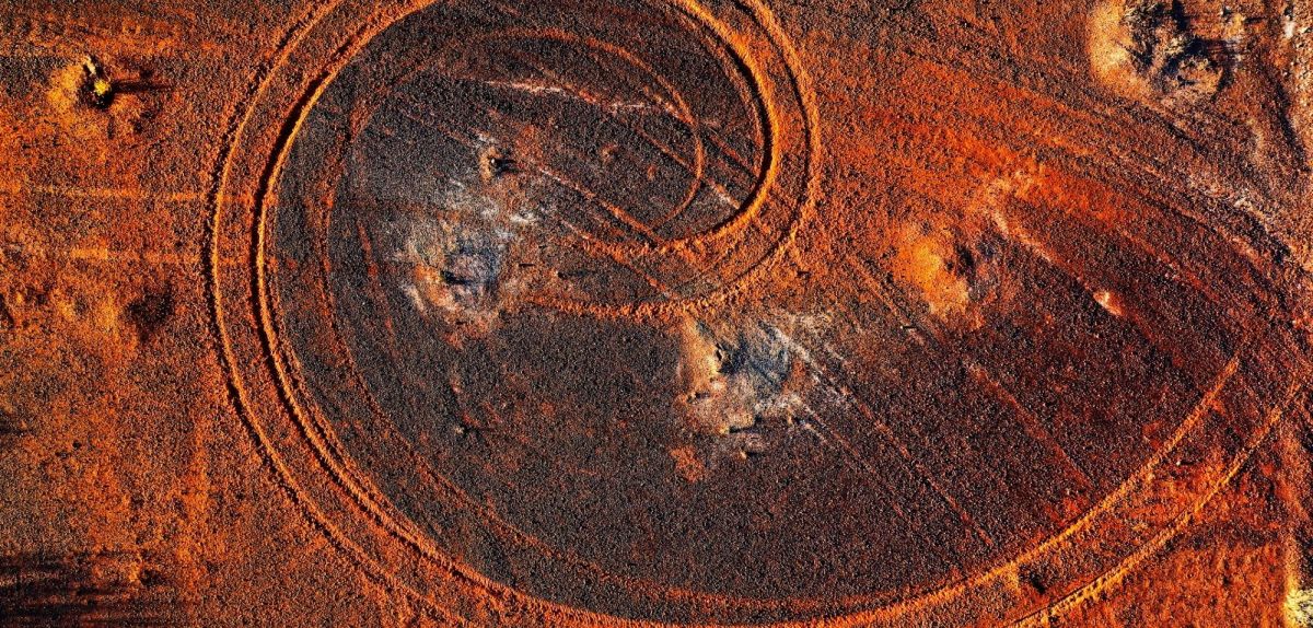 Abstraktes Wirbelmuster aus Autoreifenspuren in rotem Schmutz, beleuchtet vom Sonnenlicht der Abenddämmerung, abgelegenes ländliches Australien