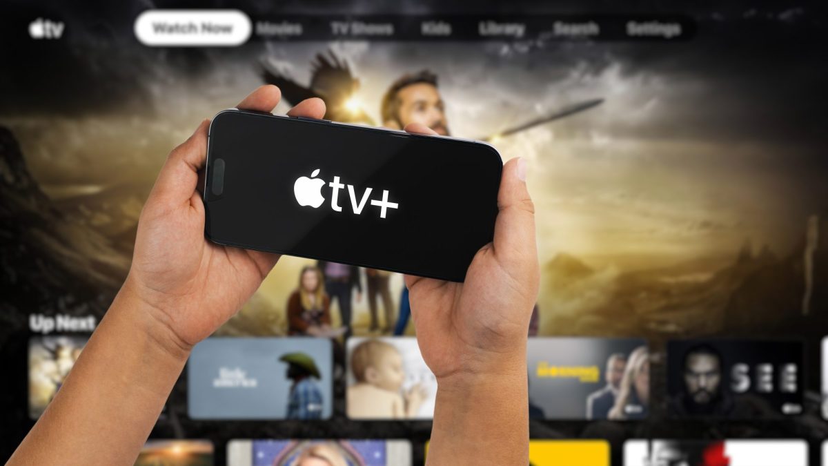 Auf einem Handydisplay wird das Apple TV+ Logo angezeigt. Im Hintergrund ist der Streaming-Dienst auf einem Fernseher geöffnet.