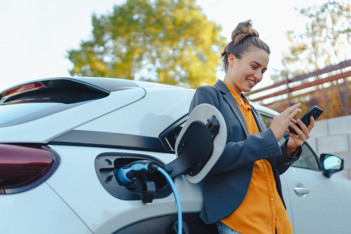 Eine Frau steht mit Smartphone an Elektroauto gelehnt, das währenddessen geladen wird.