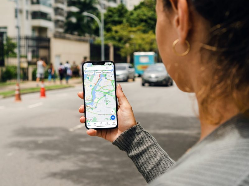 Eine Frau hält ein Handy in der Hand. auf dem Display ist die Kartenanzeige von Google Maps geöffnet. Dahinter ist eine Straße zu sehen.