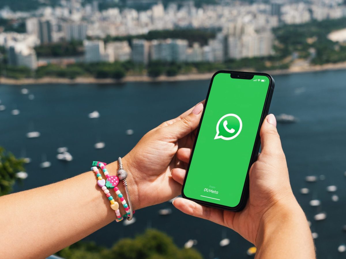 Eine Person hält ein Handy in der Hand, auf dessen Display das WhatsApp-Logo angezeigt wird. Im Hintergrund ist das Meer und die Skyline einer Stad zu sehen.