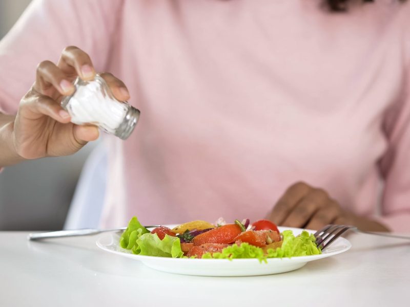 Auf einem Tisch steht ein Teller mit Essen. Eine Person hält einen Salzstreuer darüber.