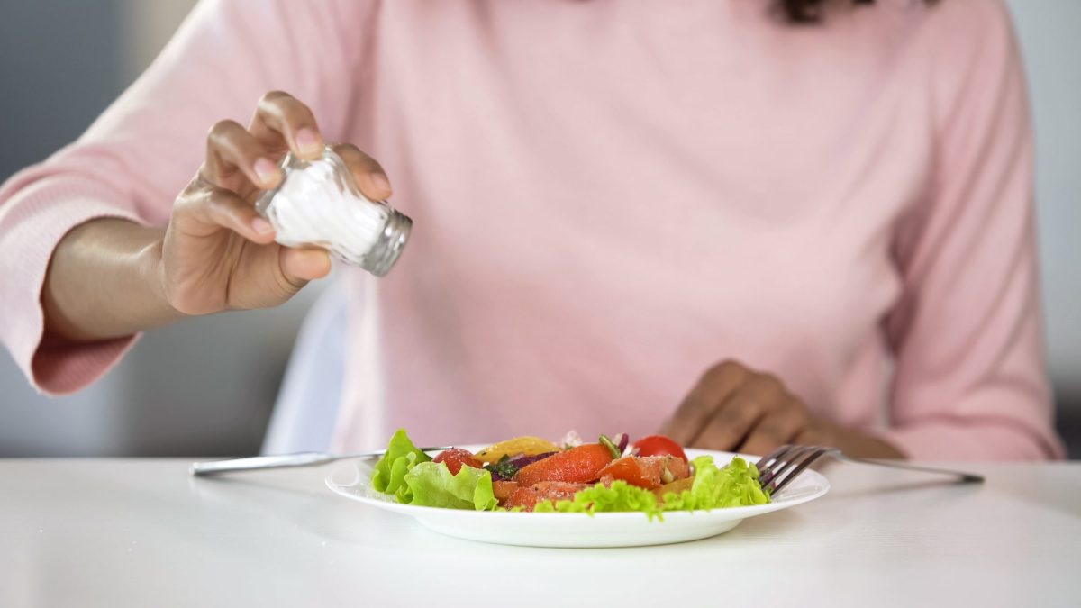 Auf einem Tisch steht ein Teller mit Essen. Eine Person hält einen Salzstreuer darüber.