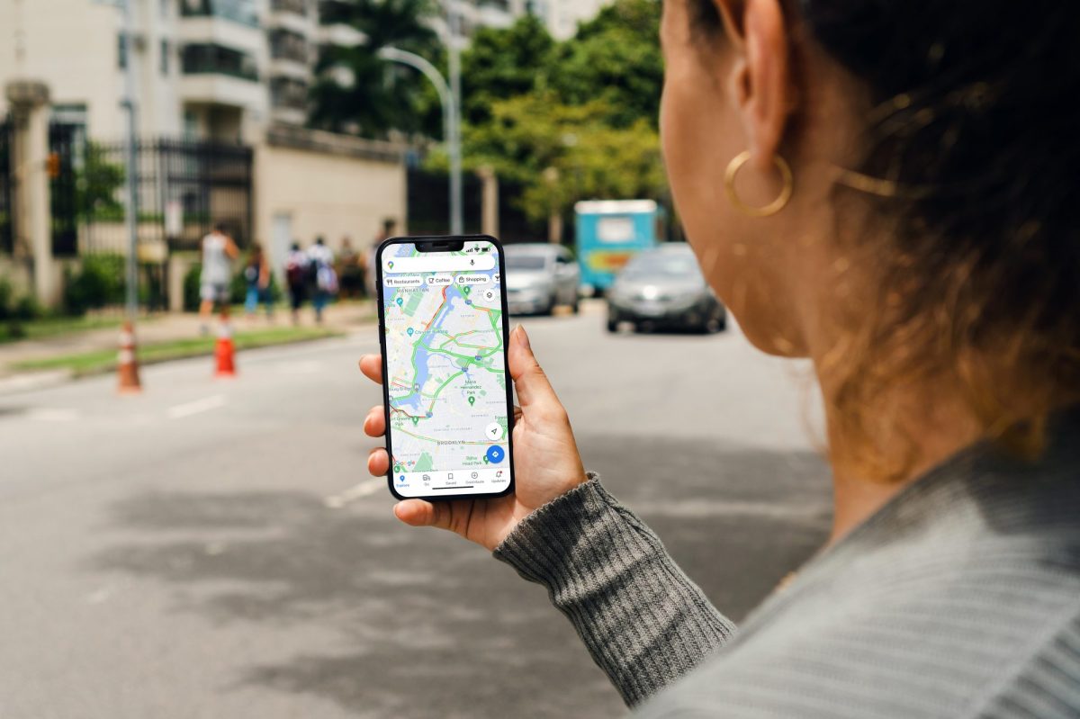 Person hält Handy in der Hand, auf dem Google Maps geöffnet ist. Im Hintergrund sind Straßen und Häuser zu sehen.