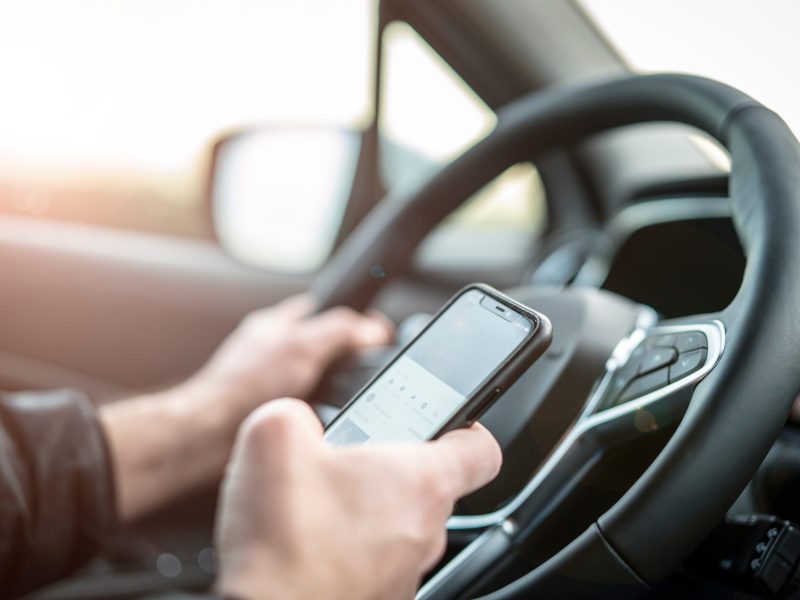 Person hält ein Handy am Steuer in der Hand.