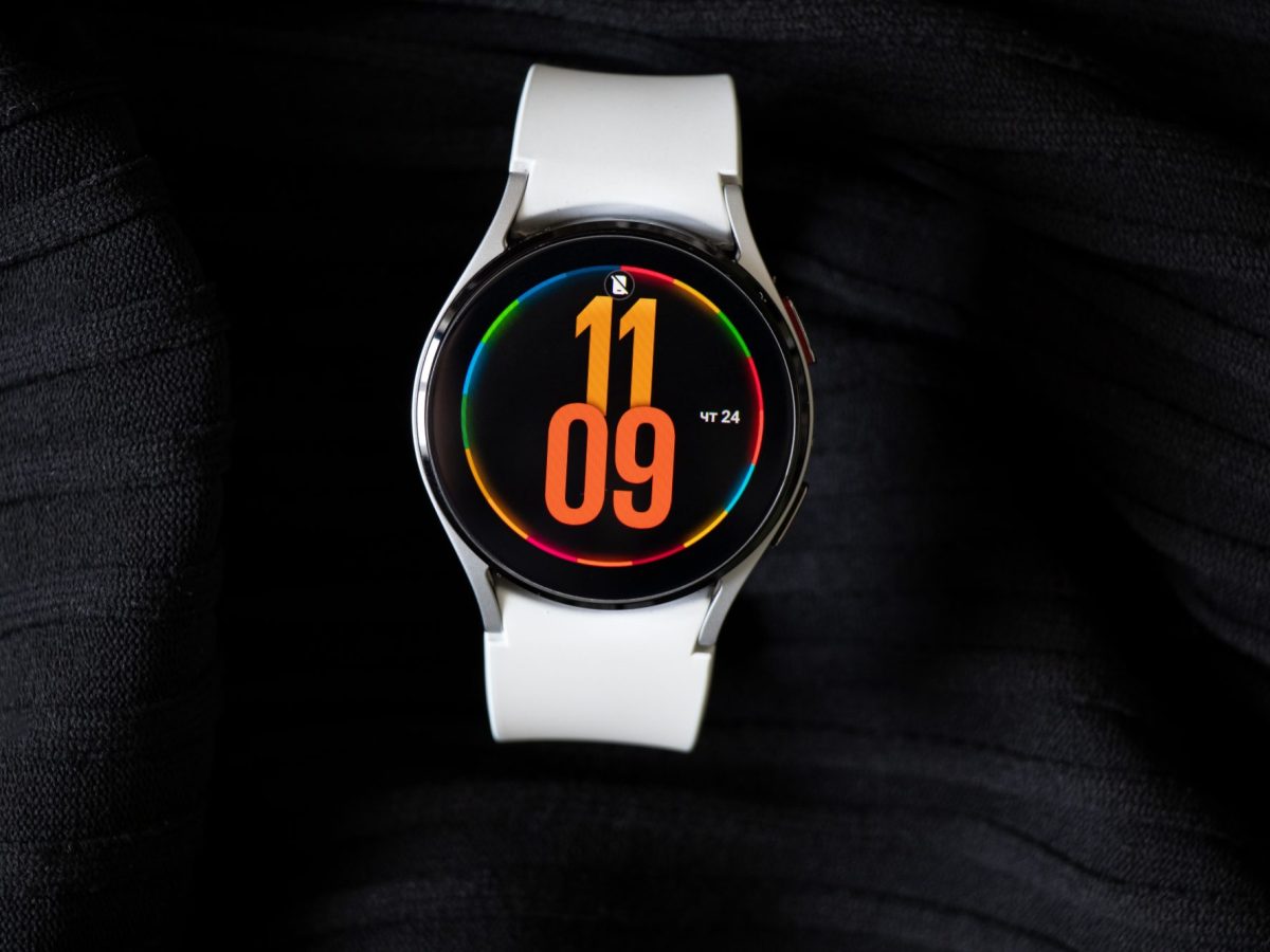 Samsung Galaxy Watch4 liegt auf einem dunklem Untergrund. Auf dem Ziffernblatt wird die Uhrzeit in oranger Schrift angezeigt.