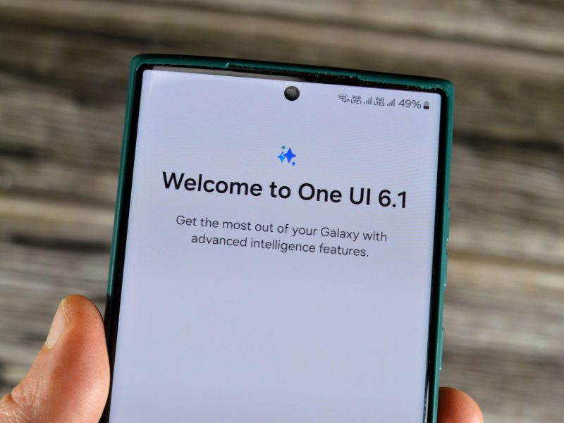 Person hält Smartphone in der Hand. Auf dem Bildschirm steht "Welcome to One UI 6.1".