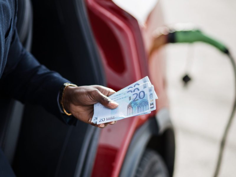 Person hält Geldscheine in der Hand. Im Hintergrund wird gerade ein Elektroauto geladen.