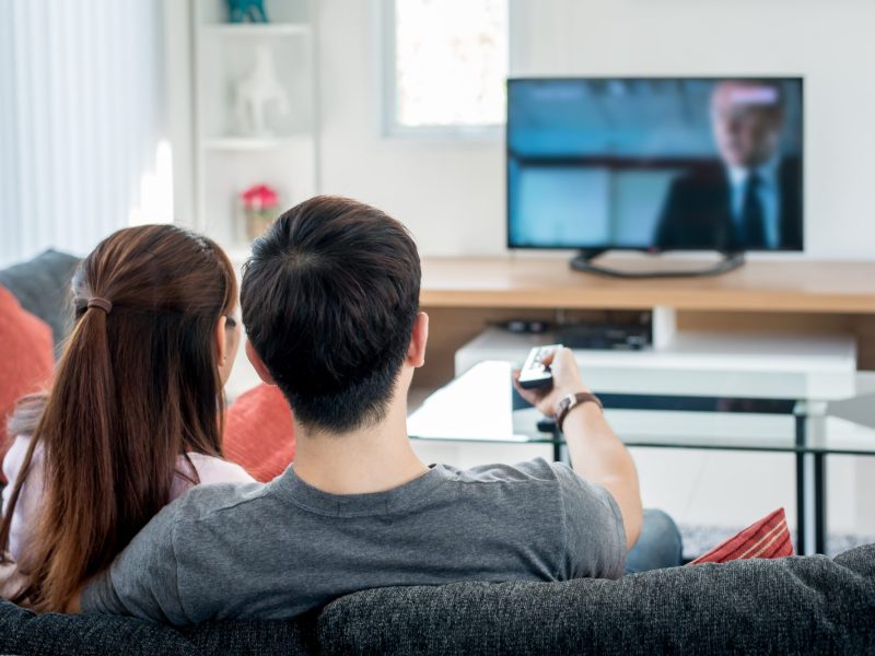 Zwei Personen sitzen auf einem Sofa und gucken auf einen Fernseher.