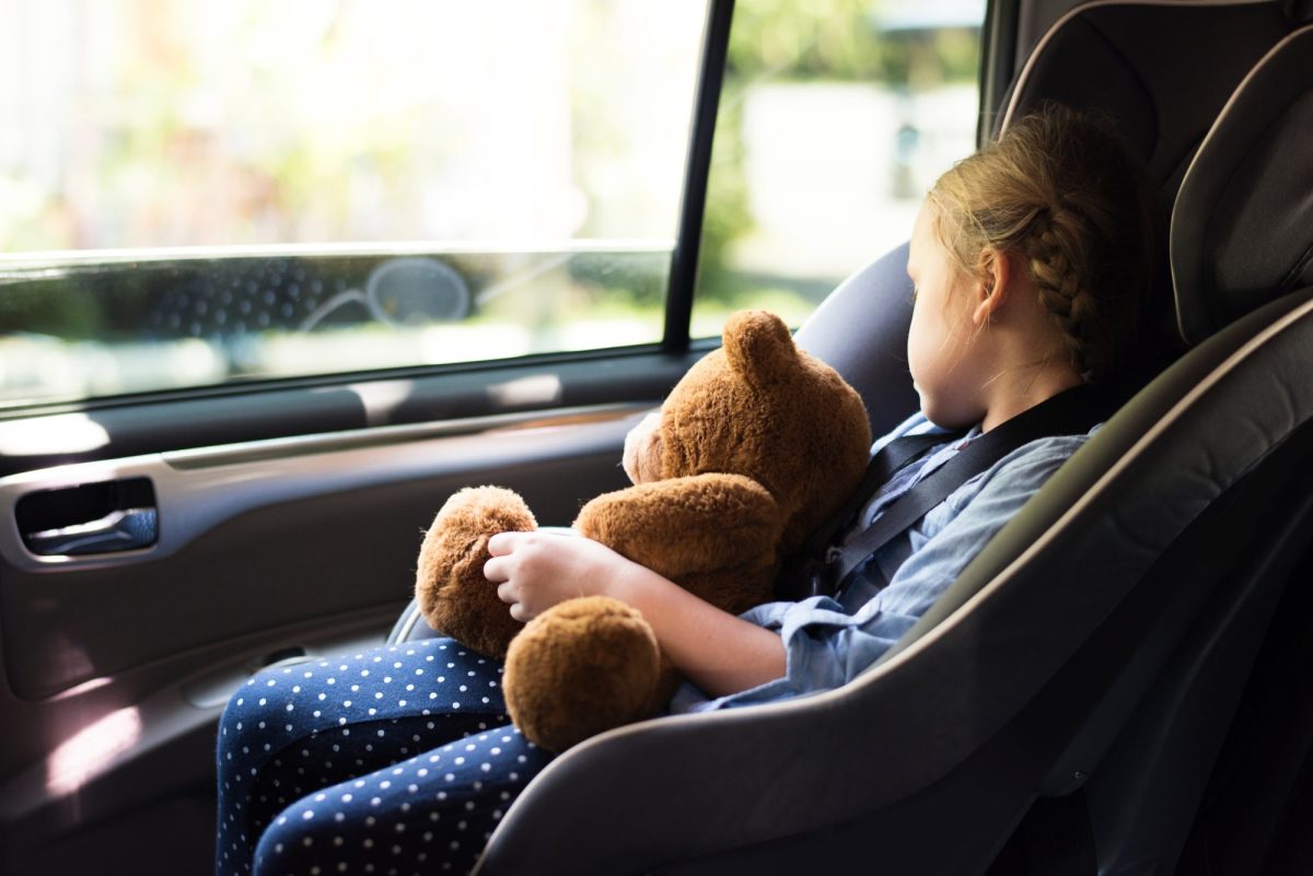 Mädchen sitzt in einem Auto-Kindersitz und hält einen Teddy in der Hand, während sie aus dem Fenster guckt.