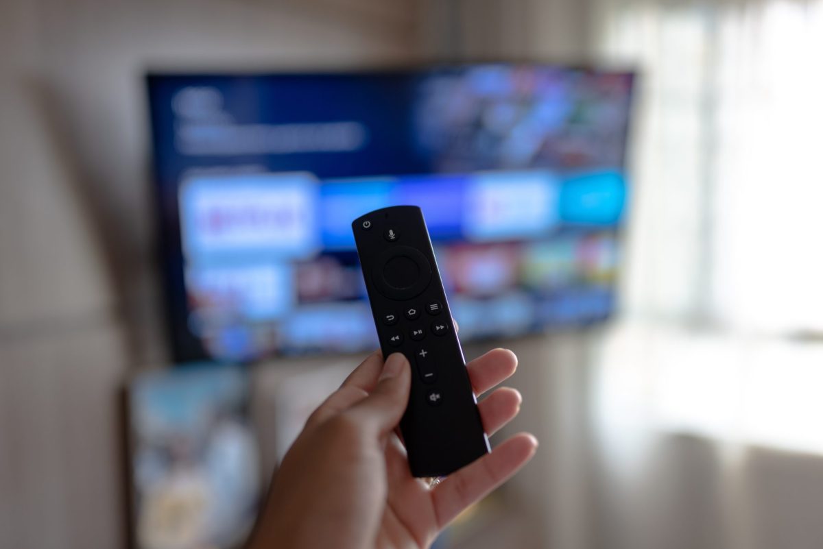 Person hält die Fernbedienung des Amazon Fire TV Sticks auf einen angeschalteten Fernseher gerichtet.