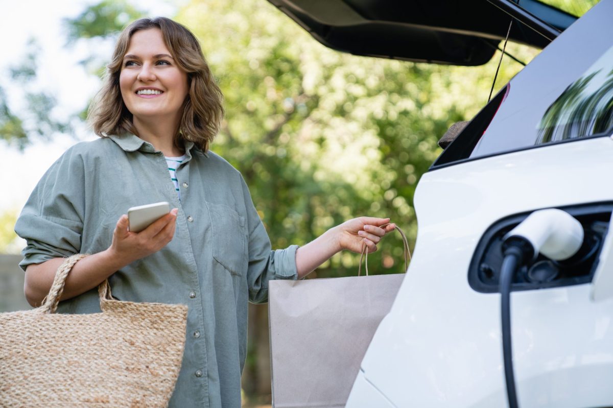 Frau lädt Einkäufe in den Kofferraum, während ihr Elektroauto geladen wird.