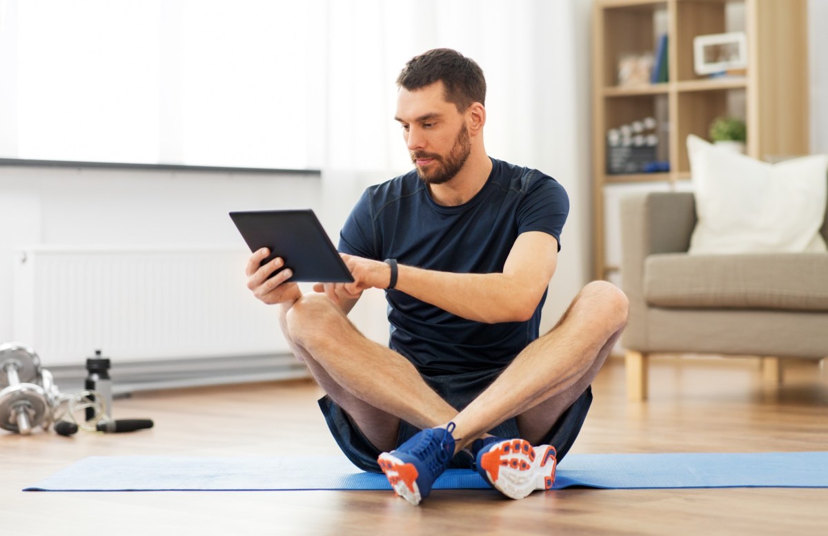 Mann schaut auf sein Tablet, während er auf einer Fitness-Matte sitzt.