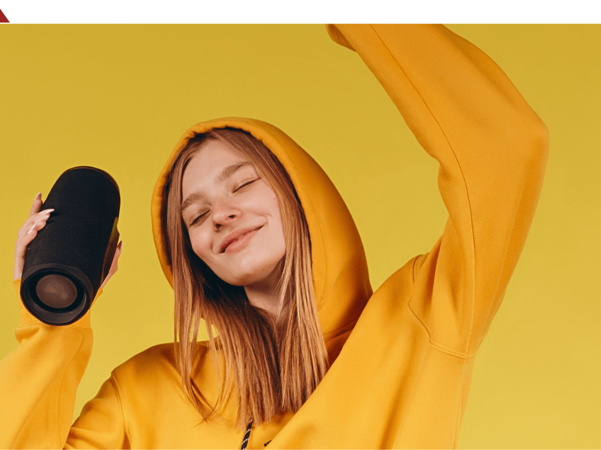 Eine Person im gelben Pullover hält einen Bluetooth-Lautsprecher in der Hand.